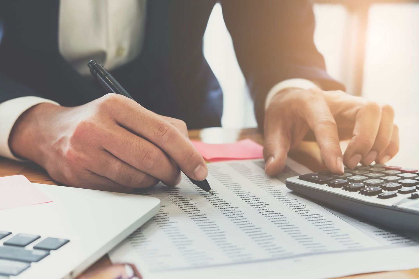 Une personne en costume examine des documents financiers avec un stylo et une calculatrice, assise à un bureau.