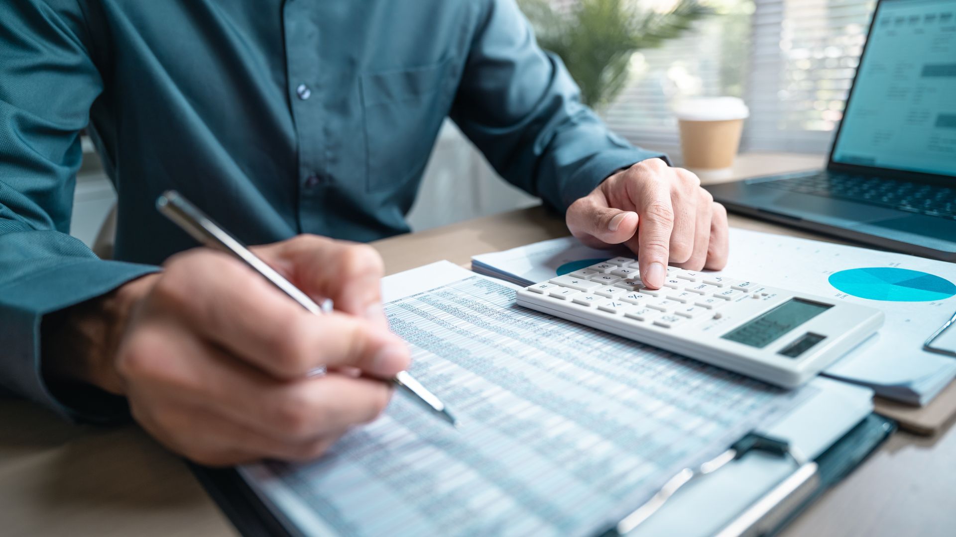 Une personne vêtue d'une chemise vert foncé travaille à un bureau, effectuant des calculs sur un document à l'aide d'une calculatrice et d'un stylo.