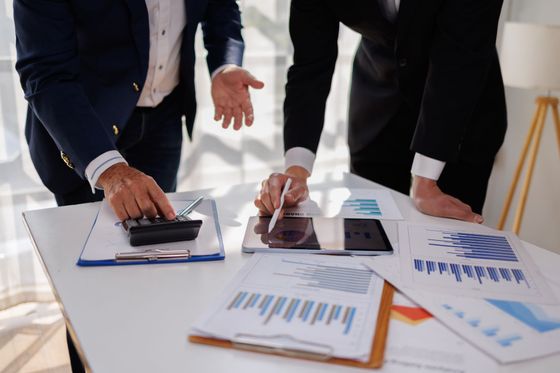 Deux professionnels en costume examinent des graphiques et des données financières sur un tableau, à l'aide d'une calculatrice et d'une tablette, dans un bureau lumineux.