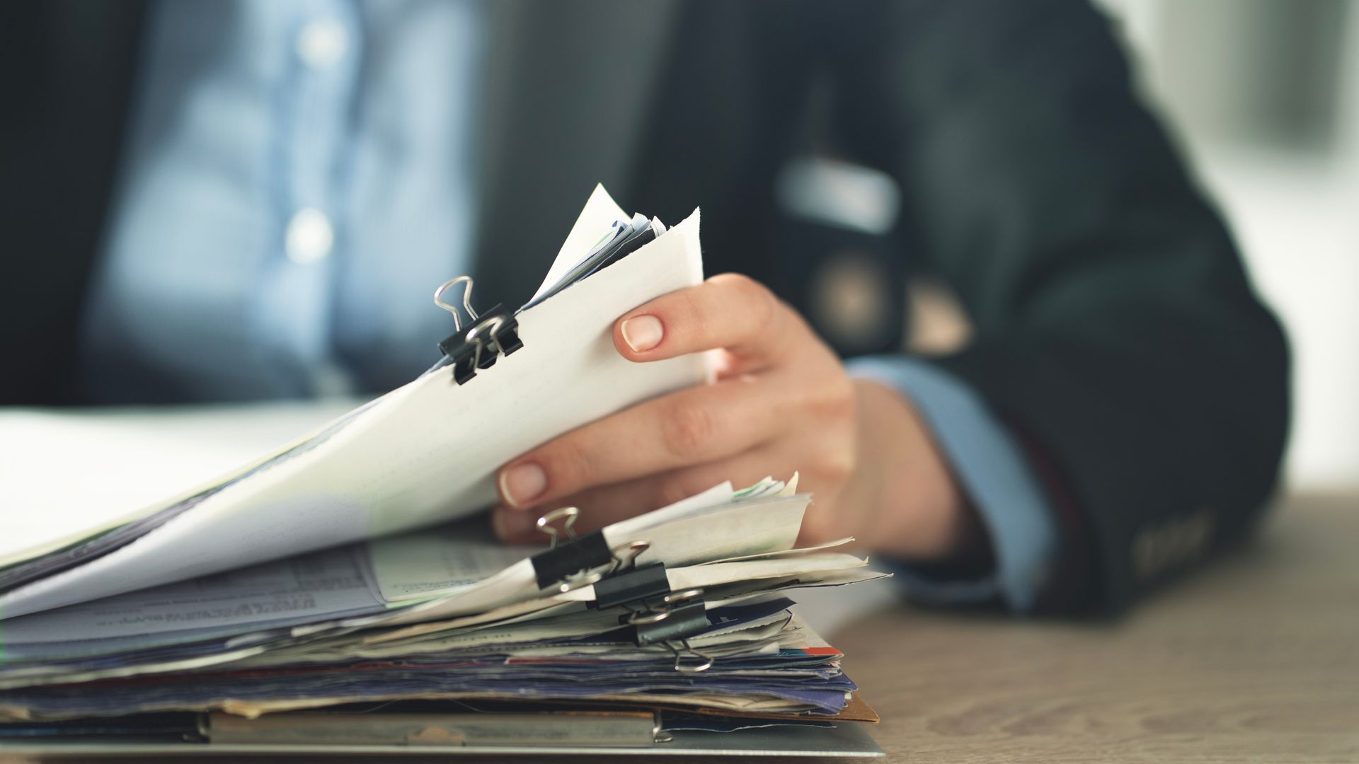 Un professionnel en costume trie une épaisse pile de papiers maintenus ensemble par des pinces à reliure sur un bureau en bois.