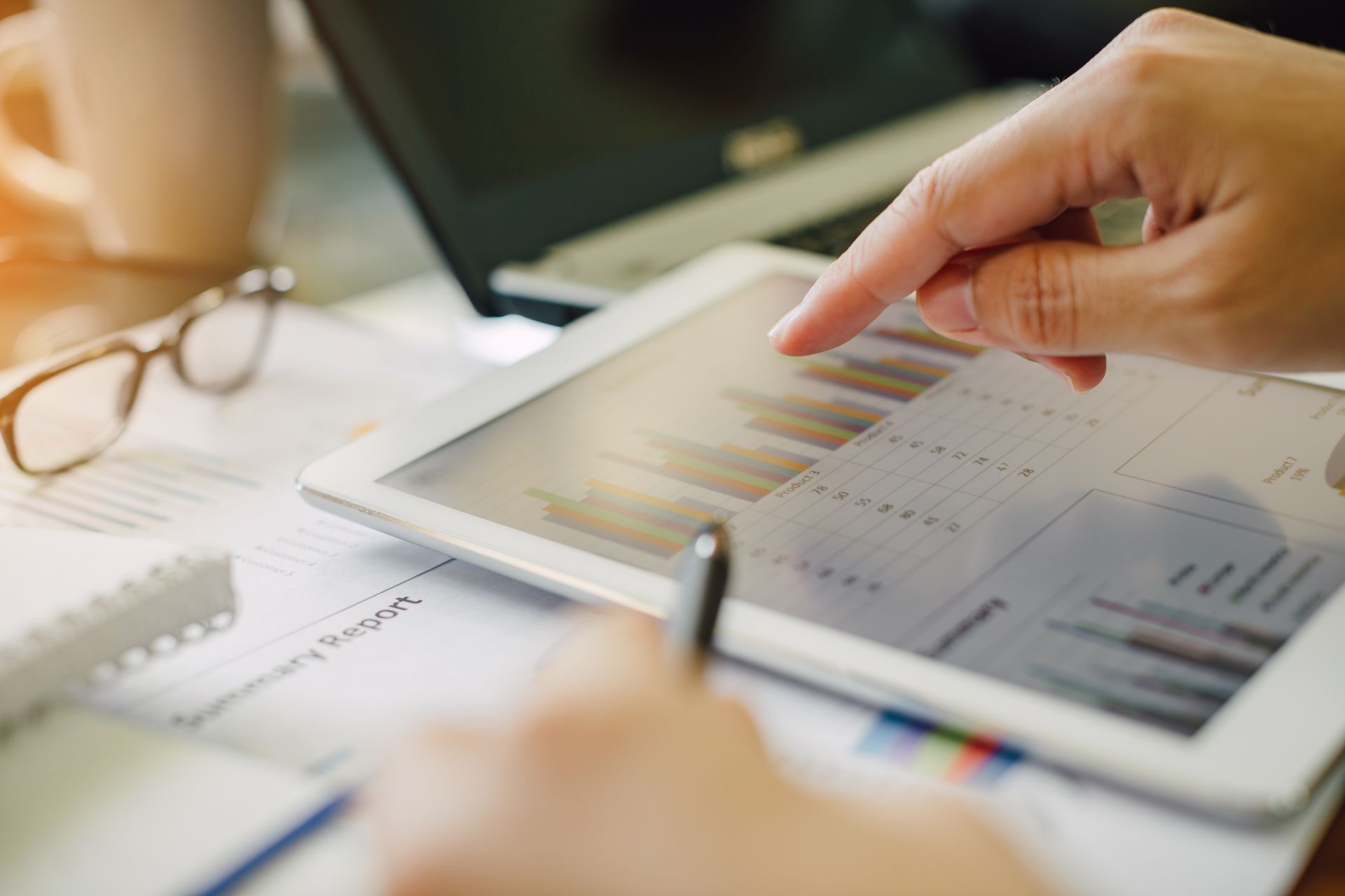 Lors d'une réunion d'affaires, une personne désigne du doigt l'écran d'une tablette affichant un graphique à barres, tandis que ses lunettes et un rapport se trouvent à proximité.