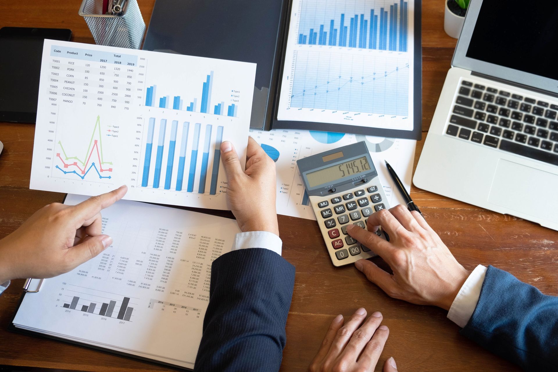 Deux personnes examinent des graphiques et des données financières sur un bureau en bois à l'aide d'une calculatrice et d'un ordinateur portable.