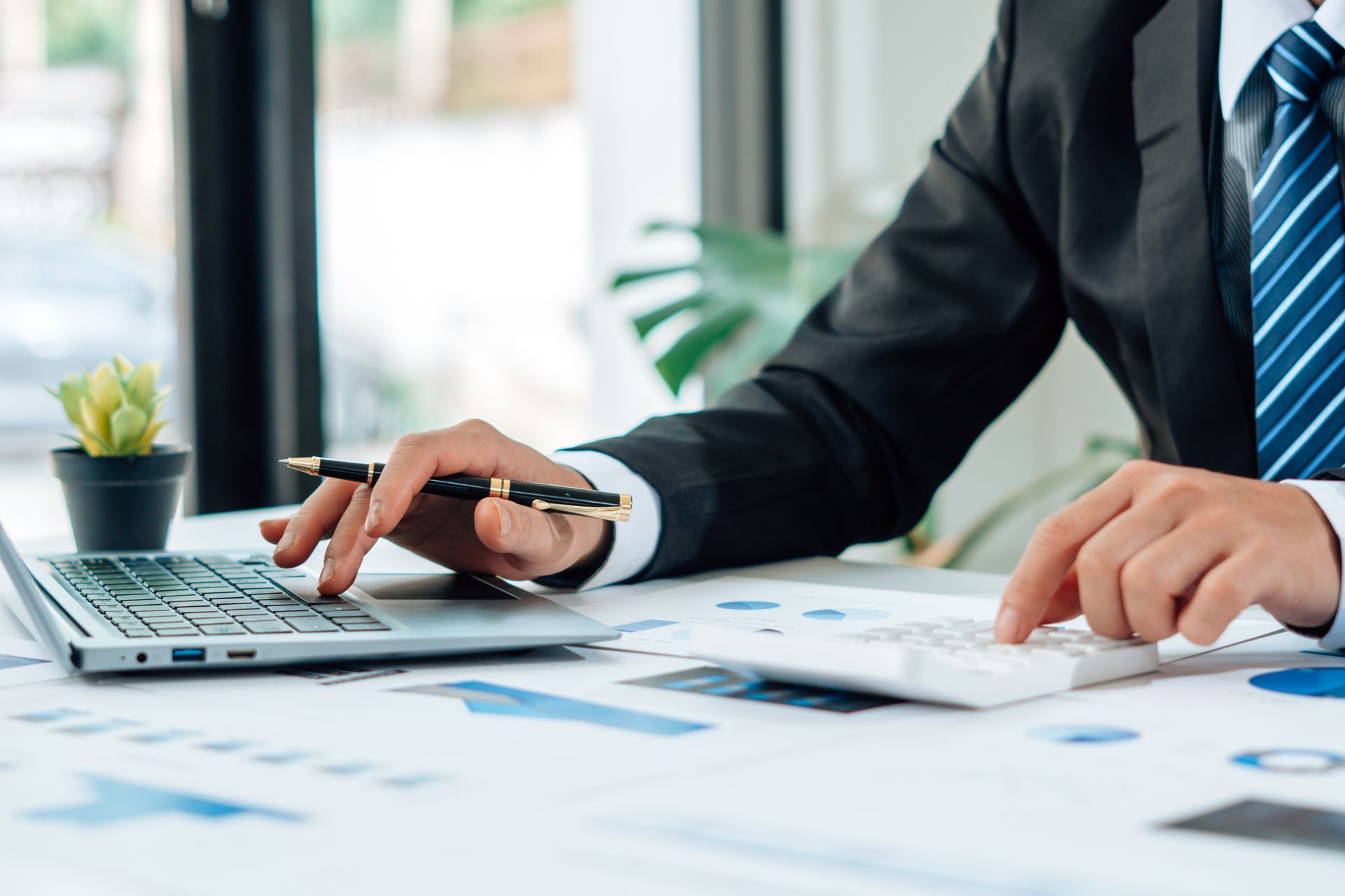 Un professionnel en costume travaille à un bureau, tapant sur un ordinateur portable et utilisant une calculatrice au milieu de divers documents commerciaux.