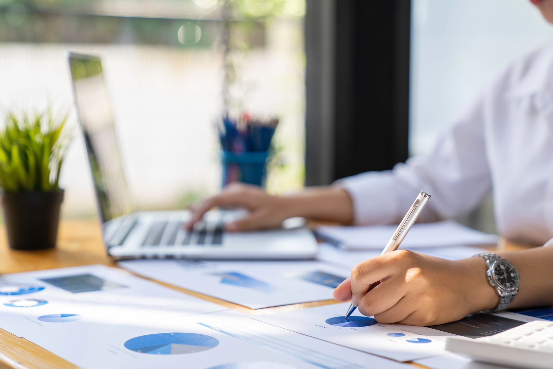 Une personne est assise à un bureau, travaillant sur un ordinateur portable tout en analysant des graphiques financiers avec un stylo.