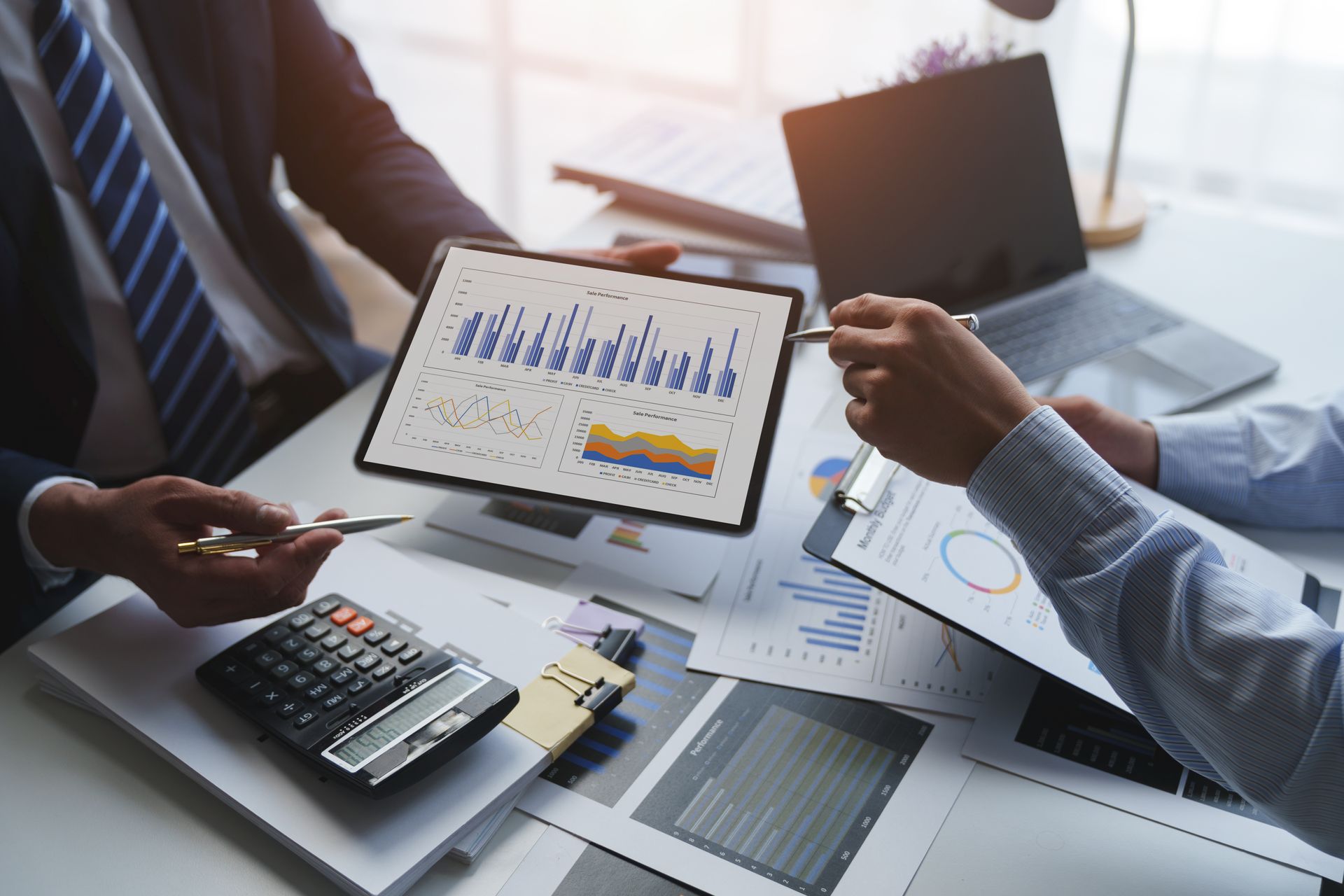Deux professionnels en tenue de ville examinent des graphiques de données financières sur une tablette numérique, assis à un bureau avec une calculatrice.