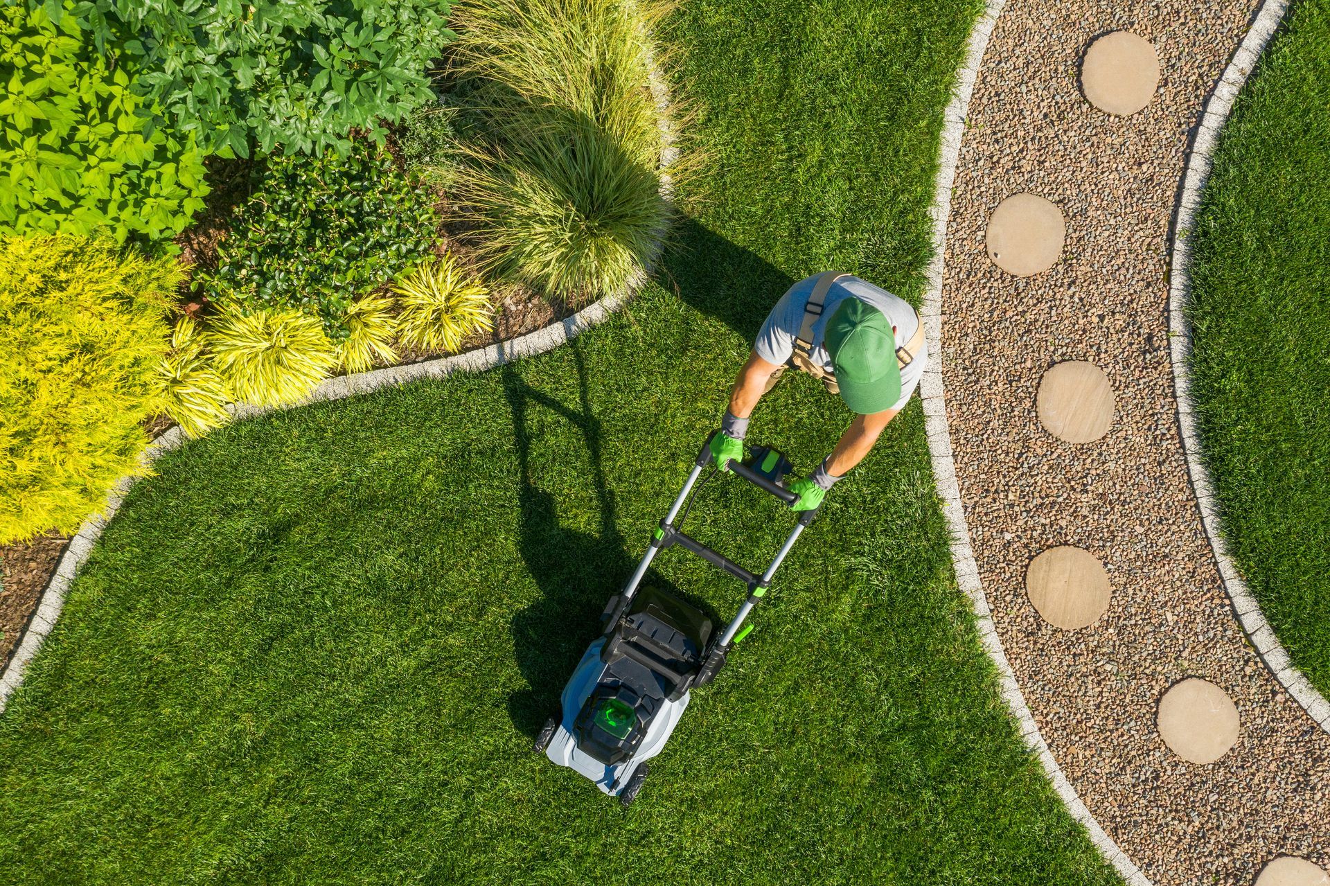 Une personne tond une pelouse verte le long d'un chemin de jardin sinueux bordé d'arbustes.