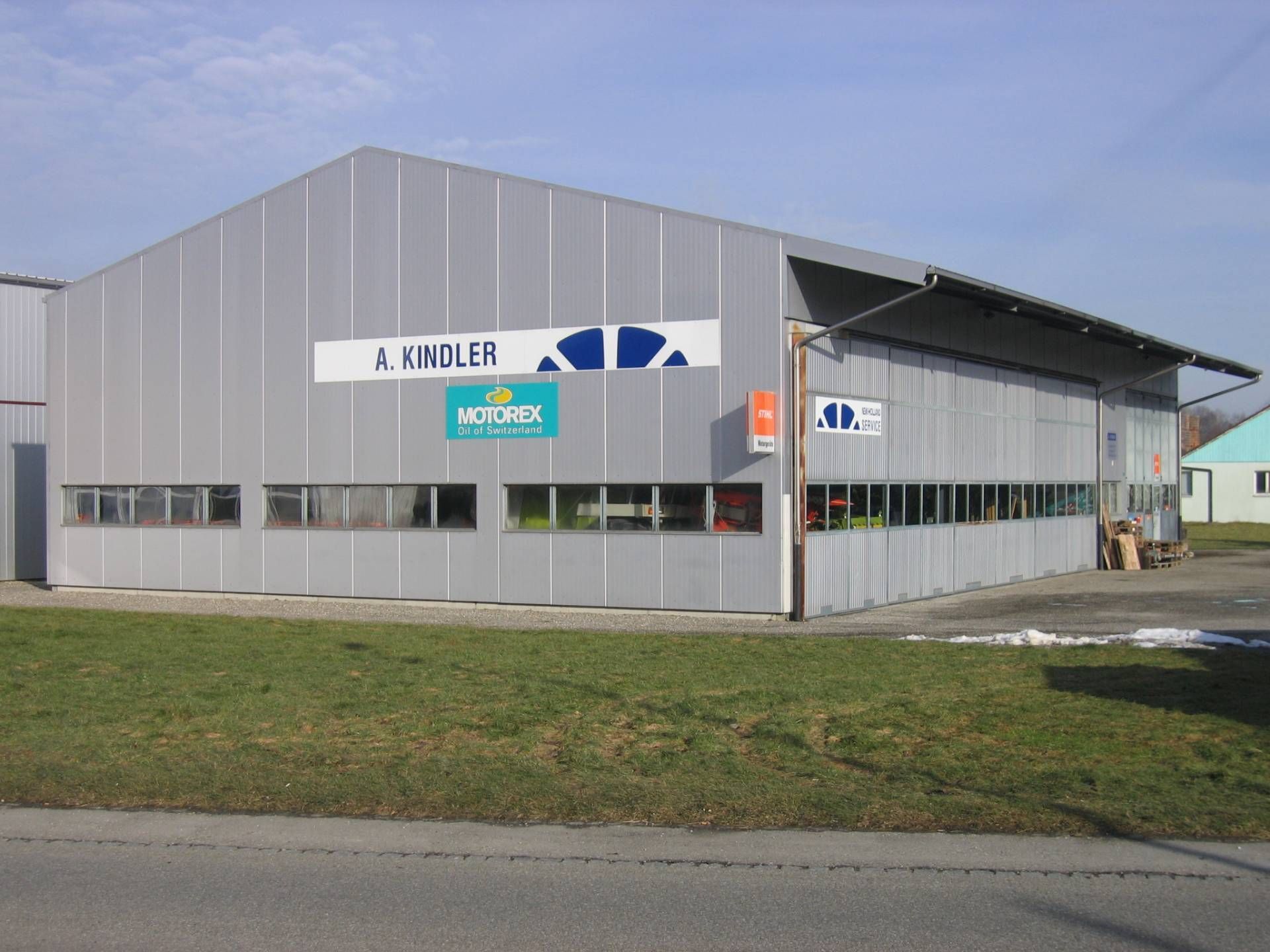 Graues Industriegebäude mit großem Tor. Die Beschilderung zeigt „A. PANDLER“ und Logos. Davor grüner Rasen.