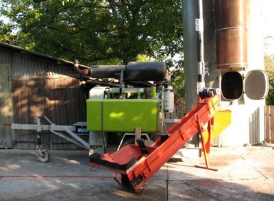 Eine grün-schwarze Maschine mit orangefarbenem Förderband steht neben einem hohen, zylindrischen Bauwerk, das in der Nähe eines Holzgebäudes geparkt ist.