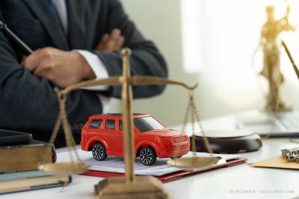 Ein rotes Spielzeugauto auf einer Waage, im Hintergrund ein Anwalt und eine Justitiastatue.