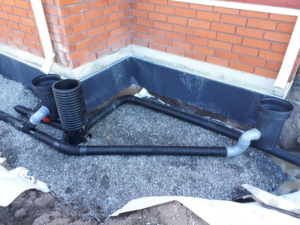 Tiilirakennuksen kulma, jossa on salaojitusputket ja tarkastuskaivot asennettuna sora-alustalle perustusten lähelle.