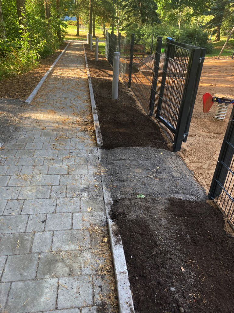 Päällystetty kävelytie kulkee tumman mullan peittämän puutarhapenkin ja hiekkatäytteisen leikkikentän reunustaman metalliaidan vieressä.