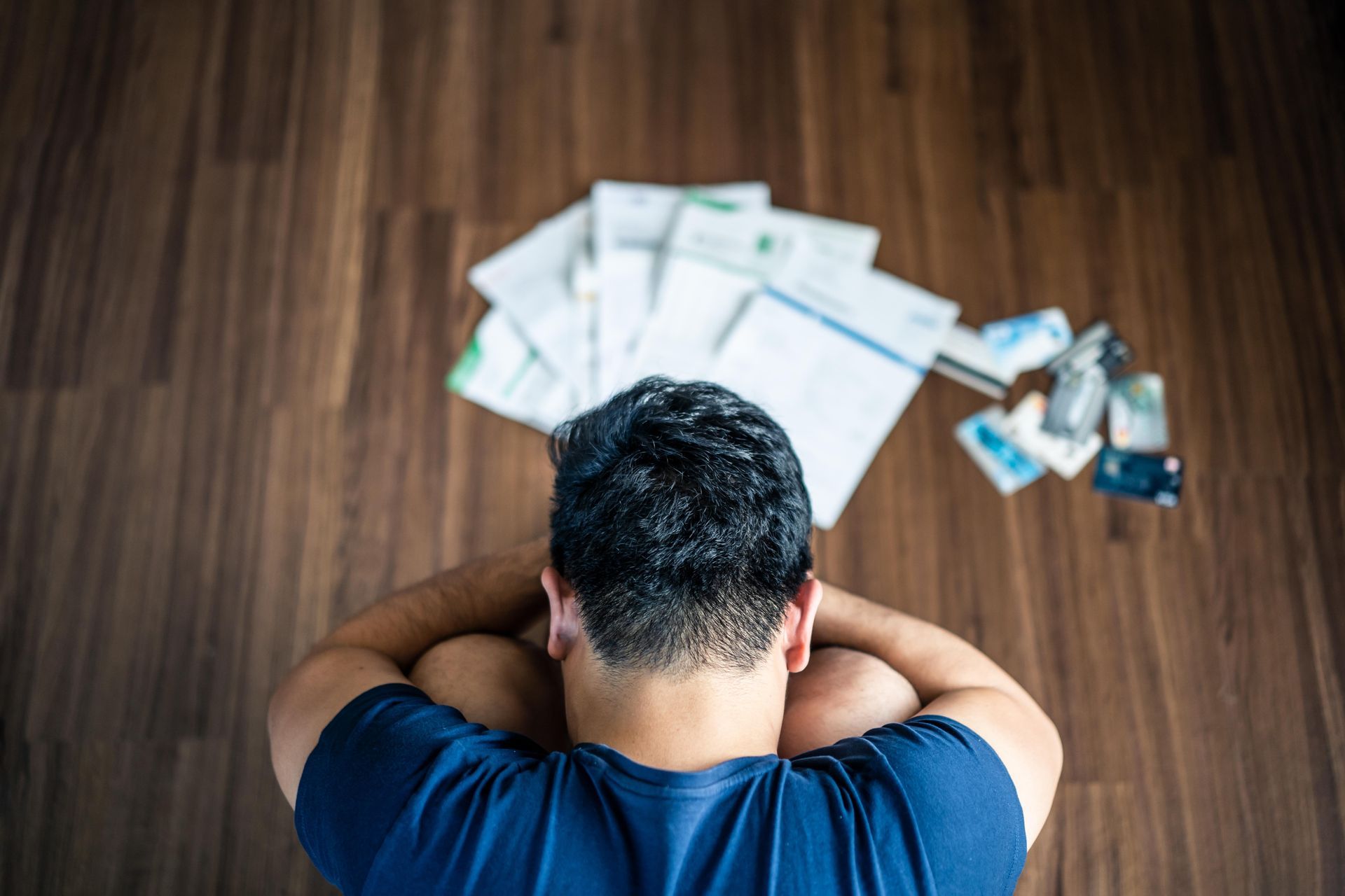 Persona con la cabeza entre las manos sentada en el suelo frente a una pila de facturas y tarjetas de crédito.