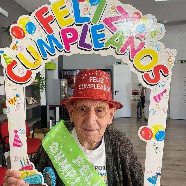 Un hombre mayor que lleva un sombrero rojo y sostiene un cartel verde que dice feliz cumpleaños