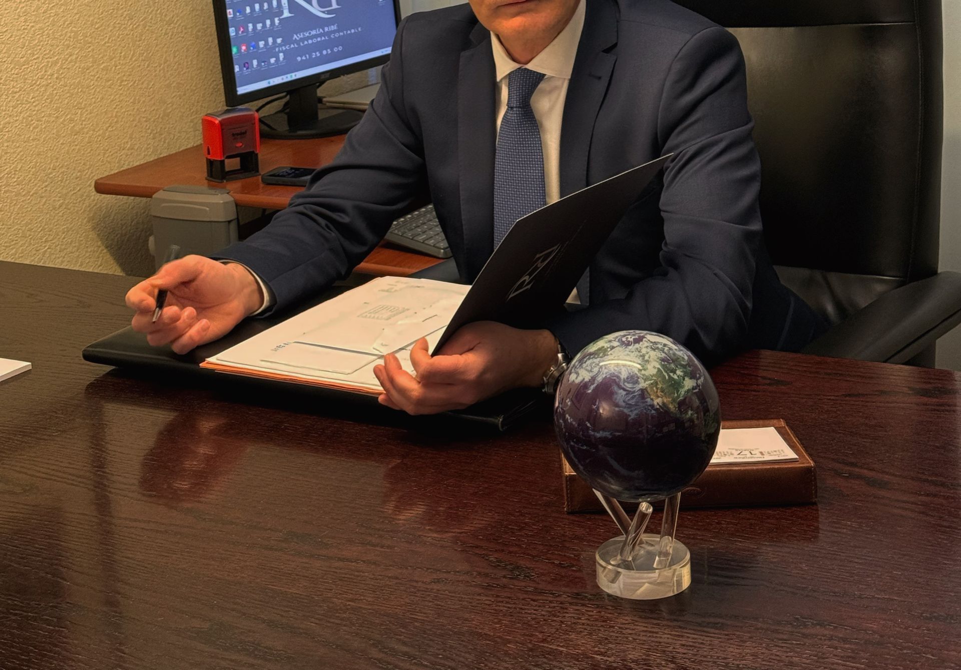 Empresario sentado en un escritorio con una carpeta y un globo terráqueo sobre la mesa, en un entorno de oficina.