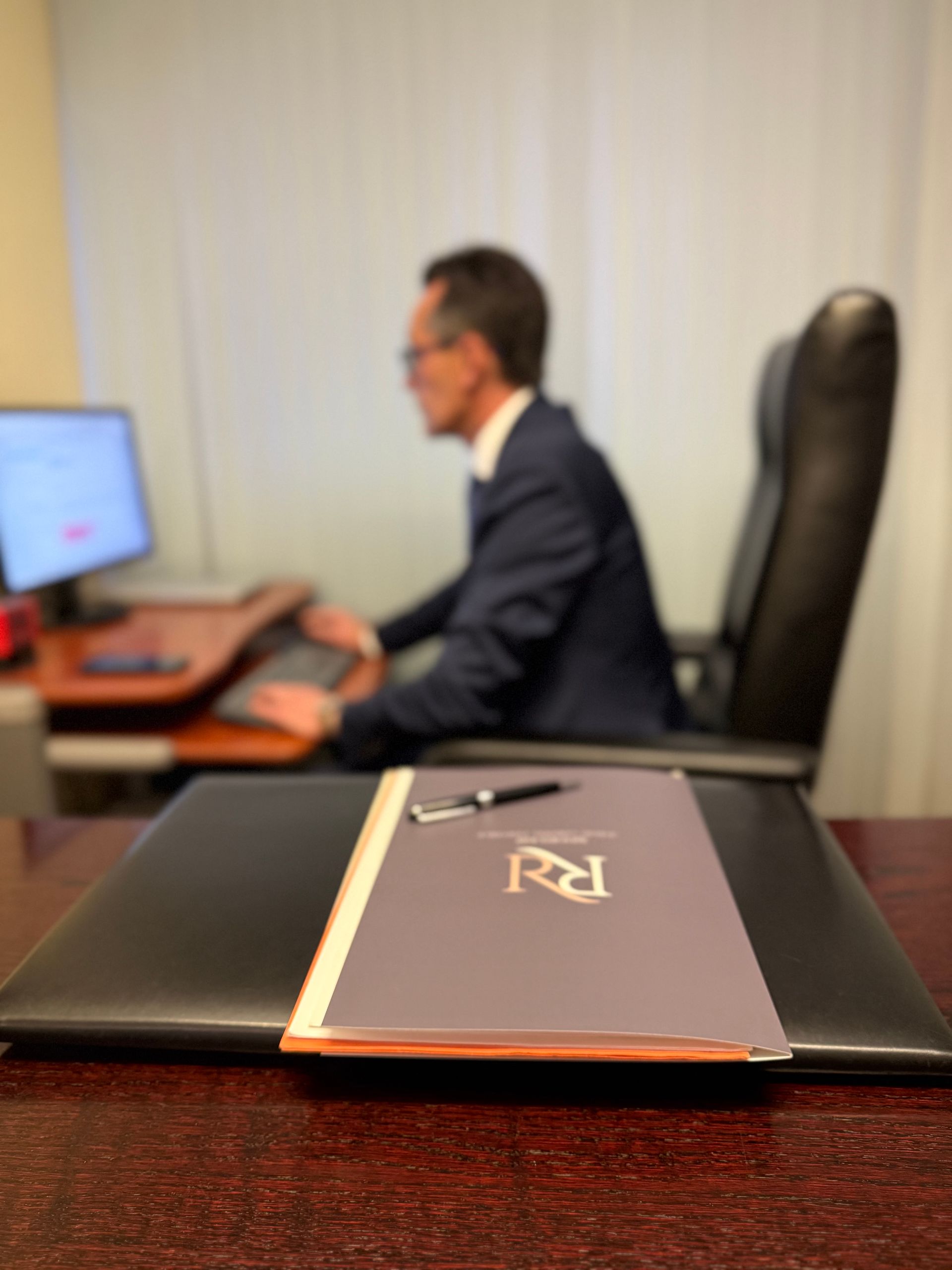 Persona sentada en un escritorio usando una computadora, con una carpeta negra en primer plano.