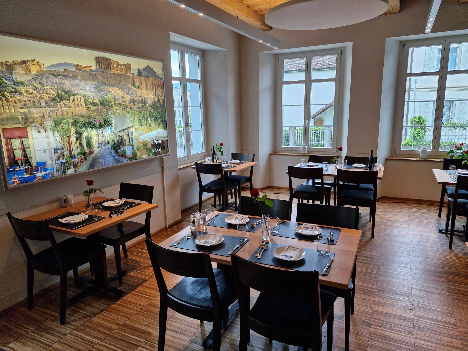 Restaurant-Speisesaal mit  gedeckten Tischen, Holzböden, grossem Bild einer griechischen Stadt an der Wand, Fenstern.