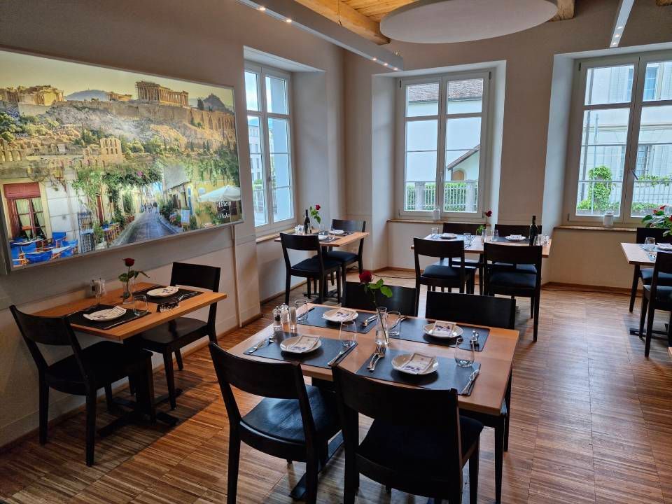 Restaurantinterieur mit zum Essen gedeckten Tischen; griechische Motive an der Wand.