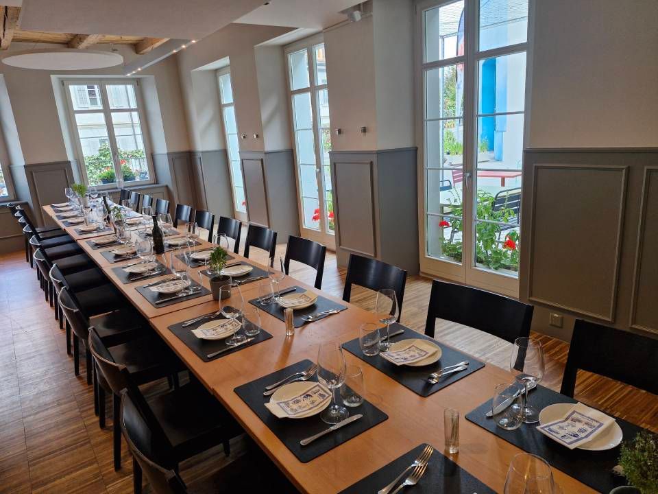 Ein langer Esstisch ist in einem hellen Raum mit grossen Fenstern für eine Mahlzeit gedeckt. Stühle stehen an beiden Seiten.