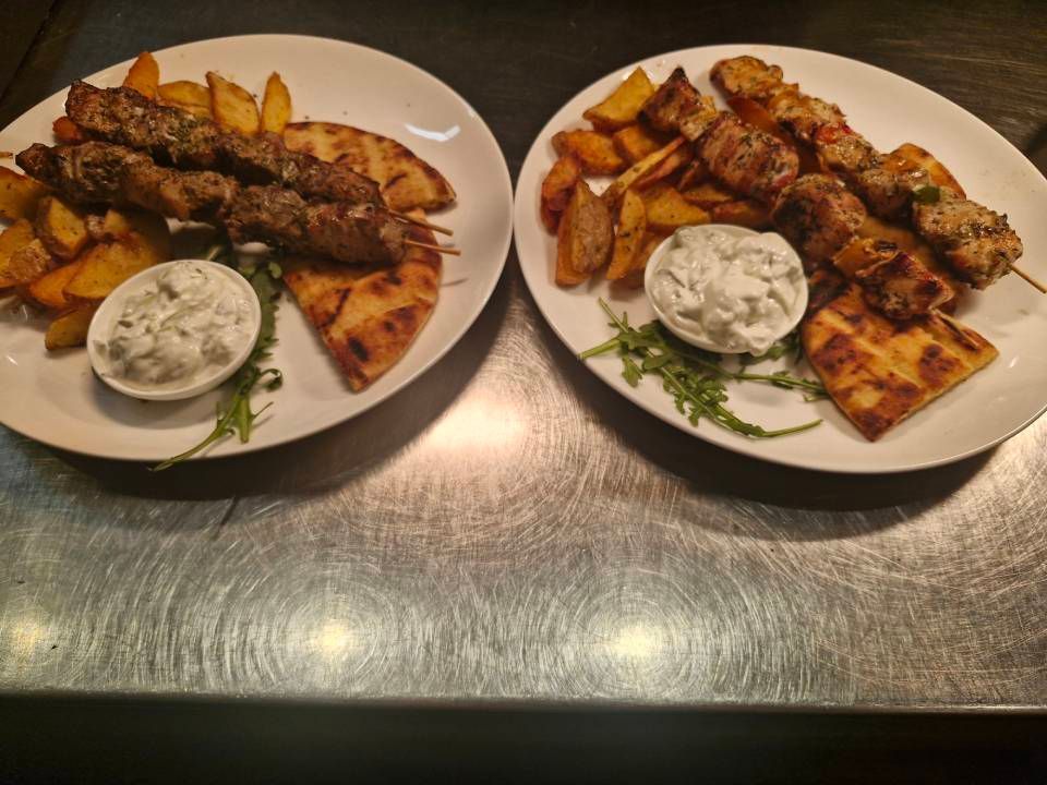 Zwei Teller griechisches Essen: Fleischspiesse, Bratkartoffeln, Pitabrot und Tzatziki-Sauce.