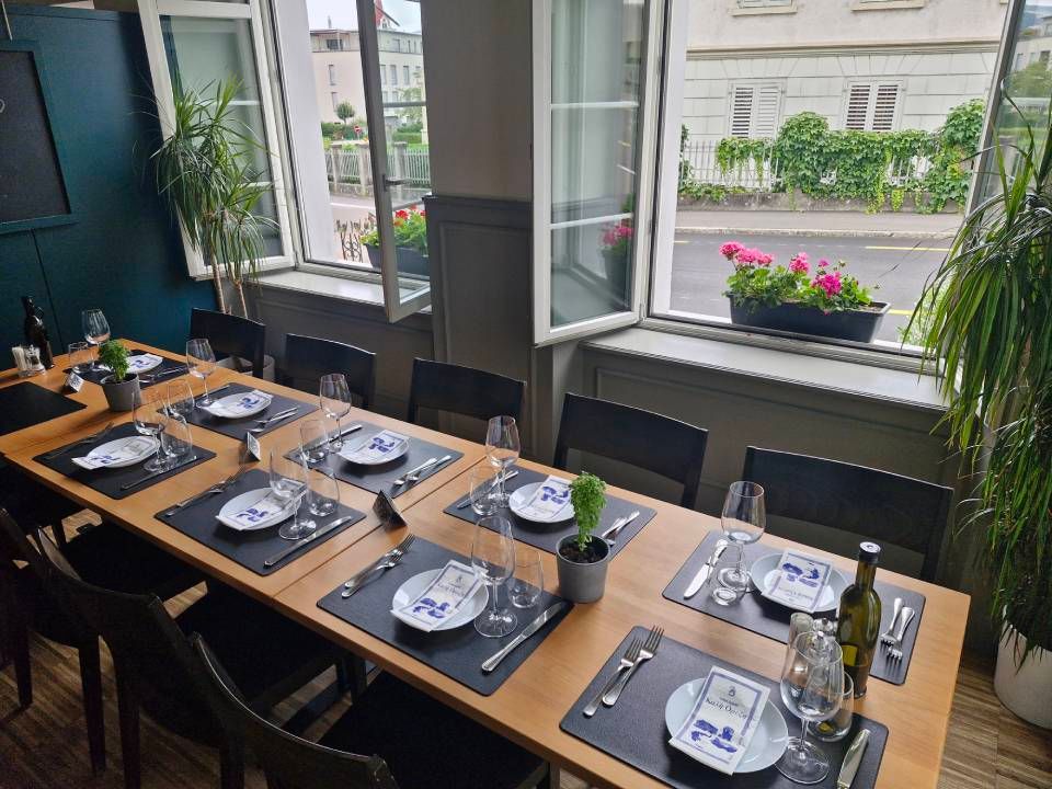 Ein Esstisch ist in einem Restaurant für eine Mahlzeit gedeckt. Die Fenster blicken auf eine blumengeschmückte Strasse.