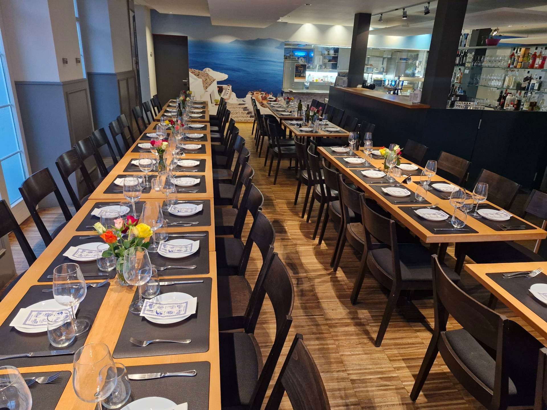 Restaurant-Speisesaal mit langen,  Tischen. Schwarze Stühle, neutrale Farbtöne und ein Wandgemälde.