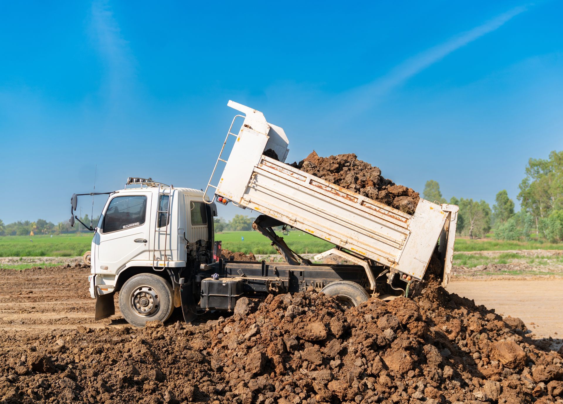 Un camion-benne blanc déverse de la terre sur un chantier boueux sous un ciel bleu azur.