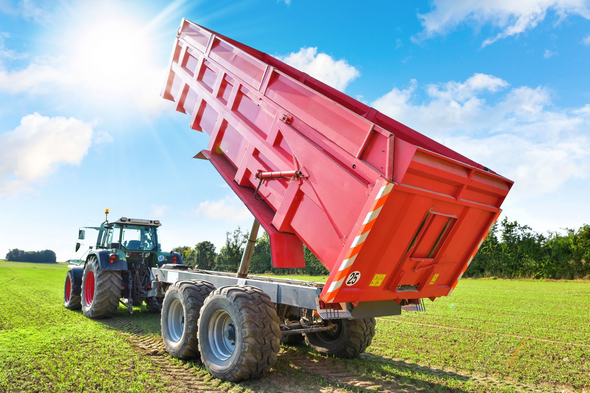 Un tracteur rouge tracte une remorque benne rouge surélevée dans un champ vert sous un ciel bleu.
