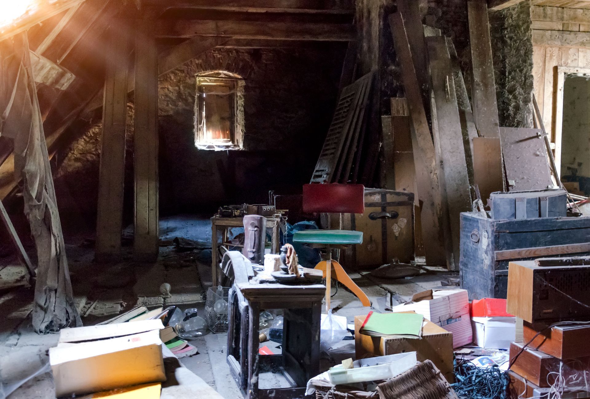 Grenier encombré de vieux meubles, de cartons et d'outils sous un toit de bois en pente, éclairé par une petite fenêtre.