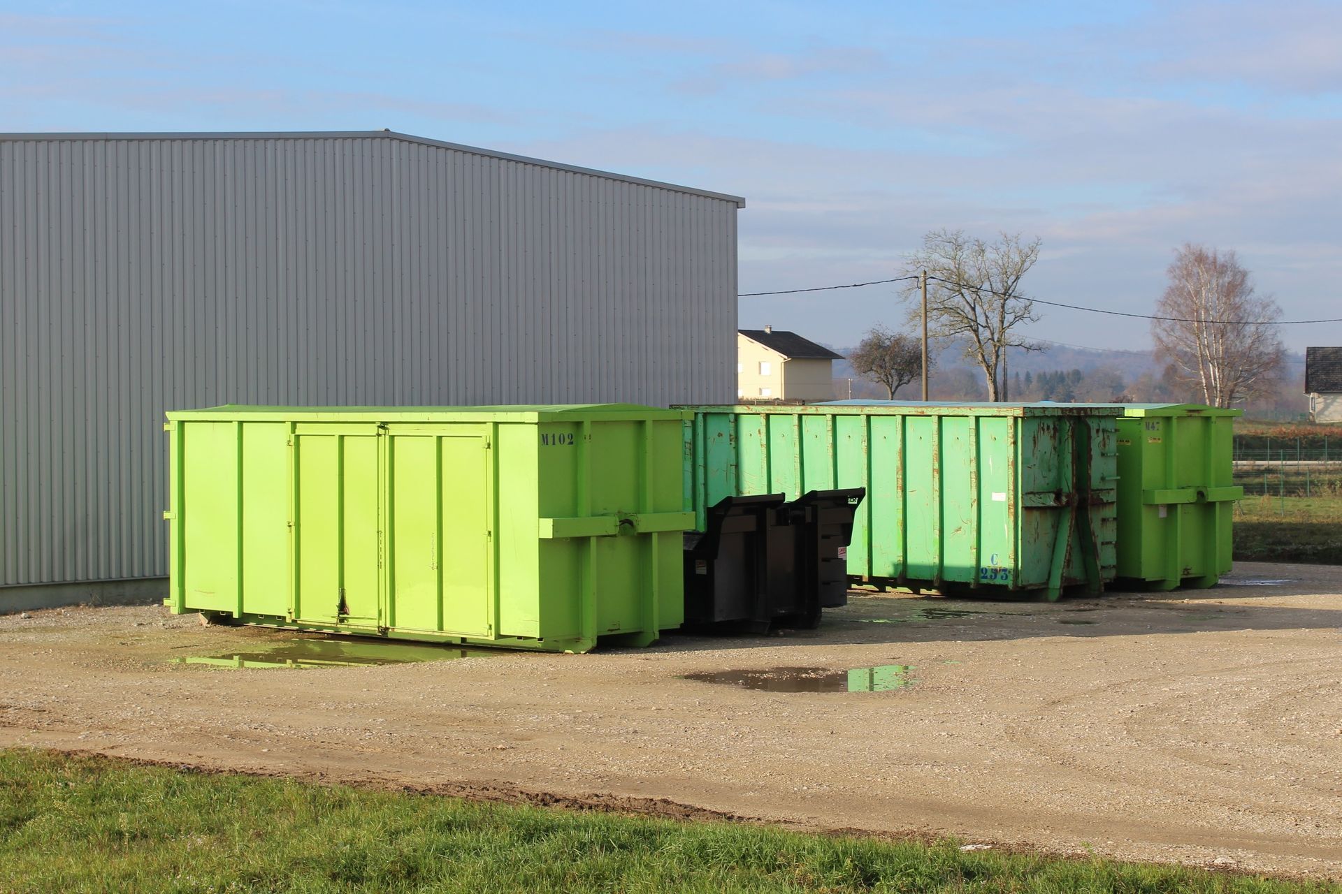 Deux bennes à ordures vert vif se trouvent à côté d'un bâtiment industriel gris sur un terrain gravillonné.