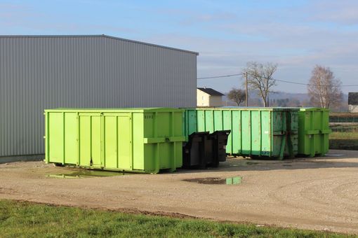 Des conteneurs à ordures verts à côté d'un grand bâtiment industriel gris sur un terrain gravillonné.