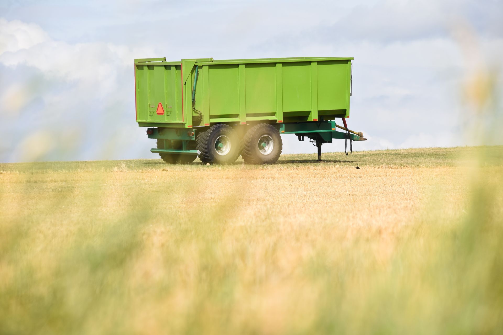 Remorque agricole verte dans un champ herbeux sous un ciel nuageux.