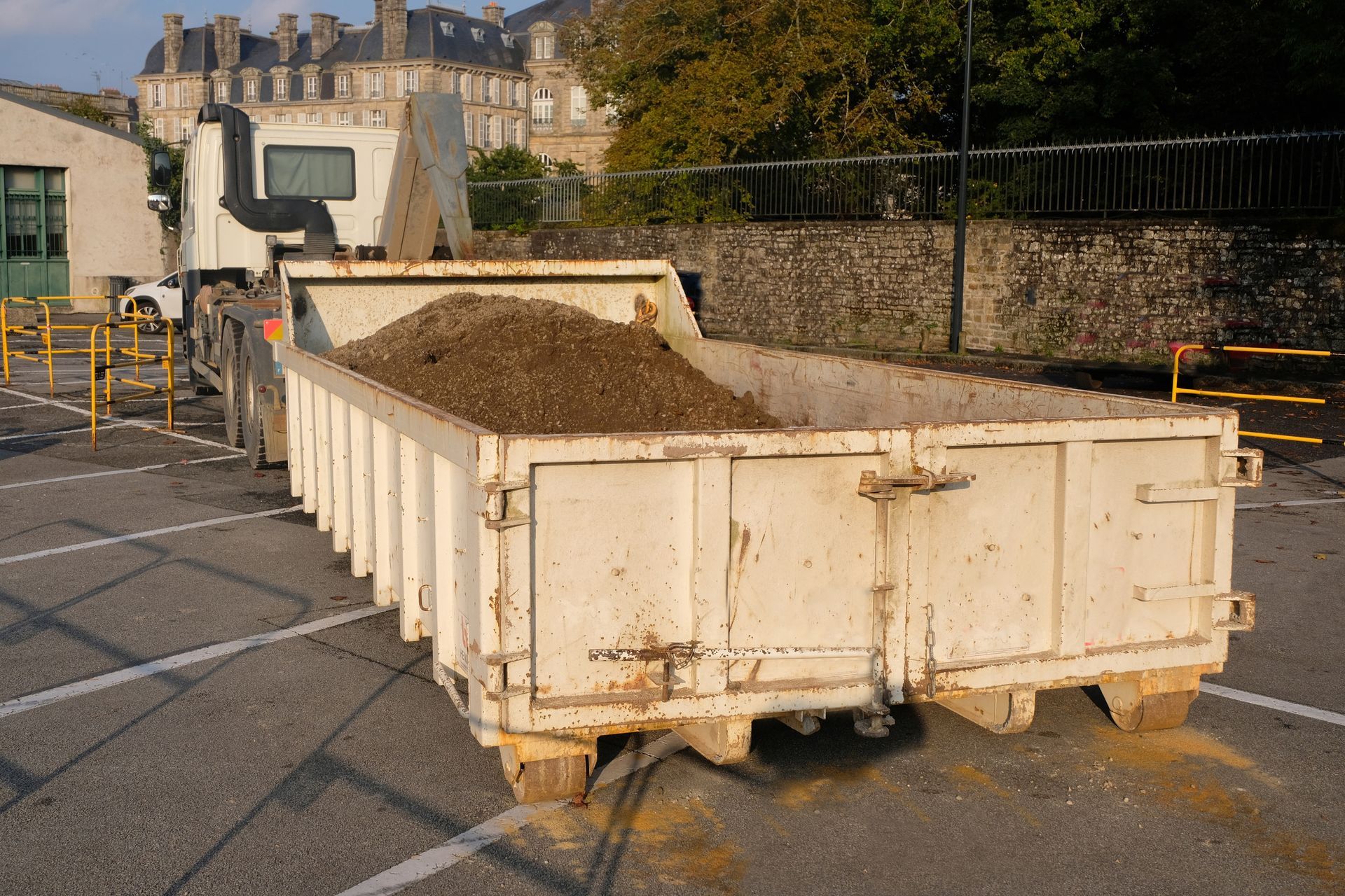 Une grande benne blanche remplie de terre se trouvait sur une rue pavée, près d'un mur de pierre et de bâtiments.