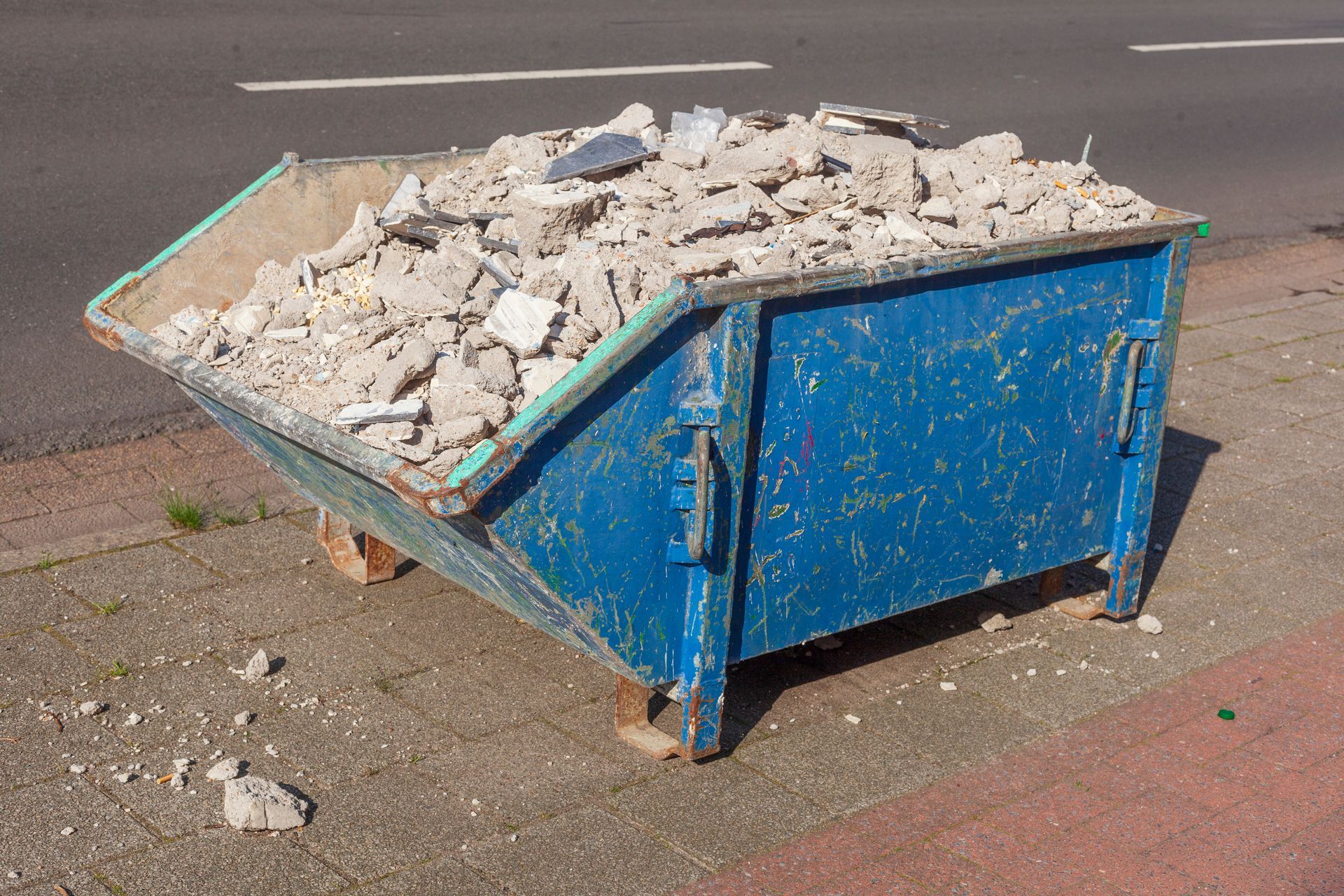 Une benne bleue débordant de gravats de béton cassés à un coin de rue pavée.