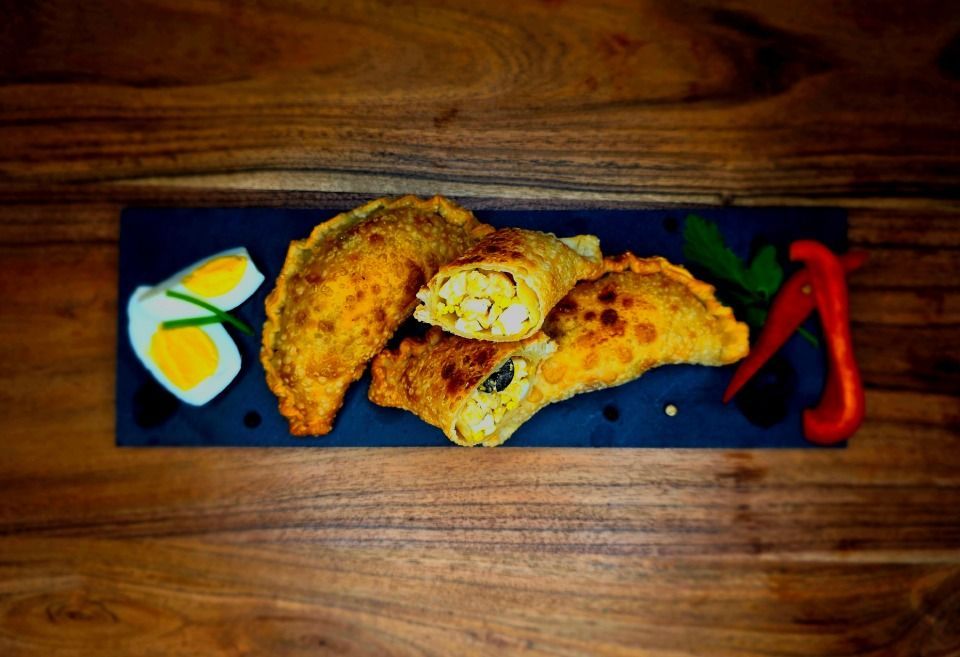 Drei frittierte Empanadas, serviert auf einem rechteckigen Schieferbrett mit in Scheiben geschnittenen gekochten Eiern, grünen Kräutern und roten Chilischoten.