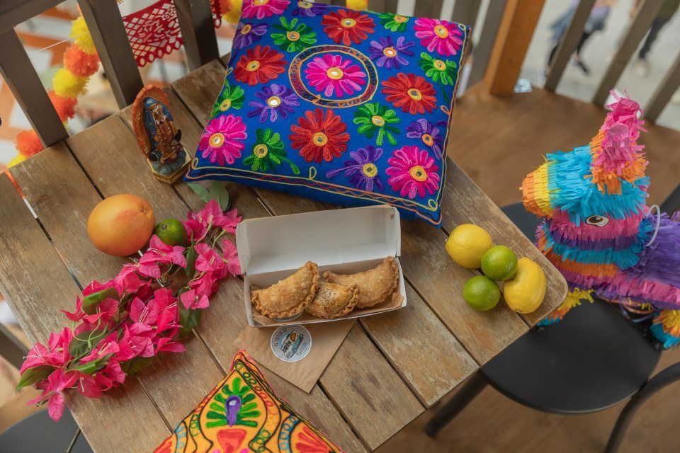 Auf einem Holztisch stehen eine Schachtel Empanadas, bunte Kissen mit Blumenmuster, eine kleine Piñata, Zitrusfrüchte und leuchtend rosa Blumen.