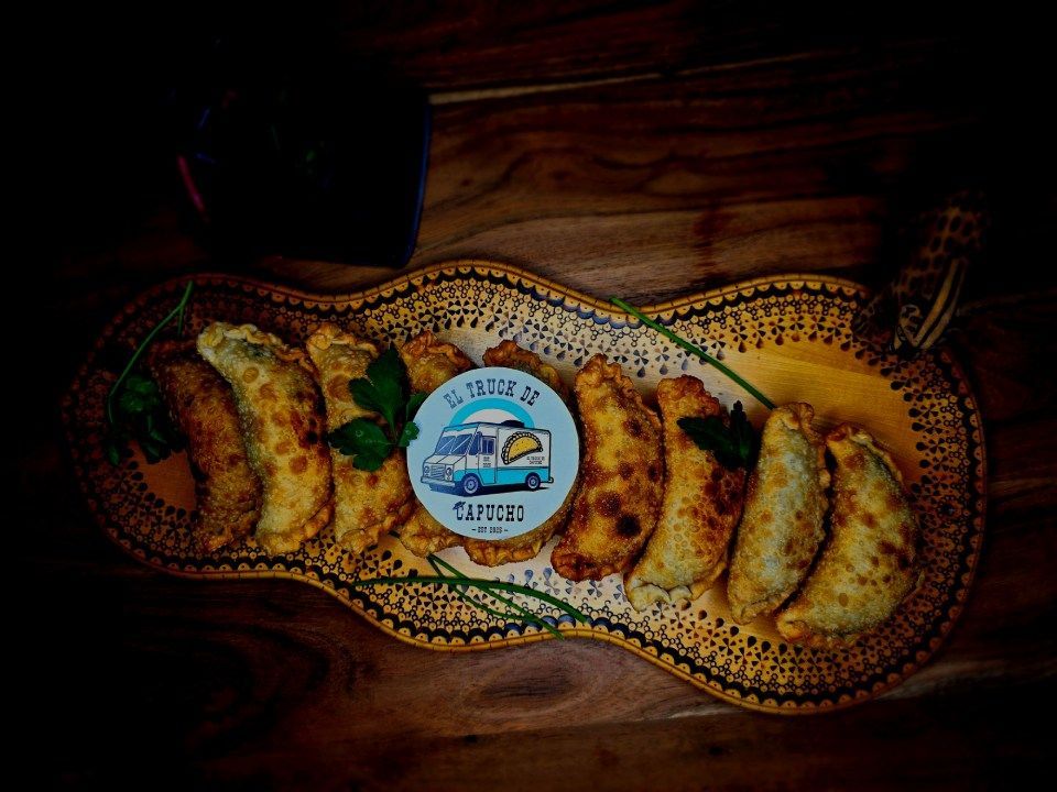 Auf einer dekorativen Platte aus Holz liegen acht goldbraune Empanadas mit einem runden Markenlogo in der Mitte.