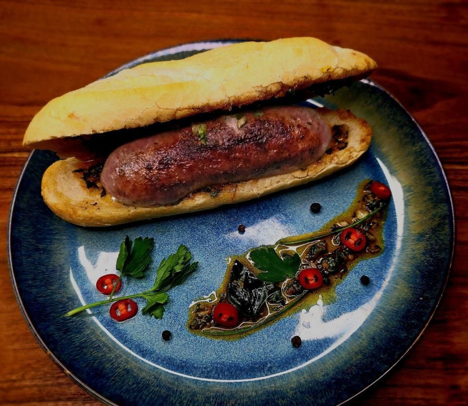 Eine gegrillte Wurst in einem gerösteten Brötchen auf einem blauen Keramikteller, garniert mit einem Klecks Kräutersauce, Paprika und Petersilie.