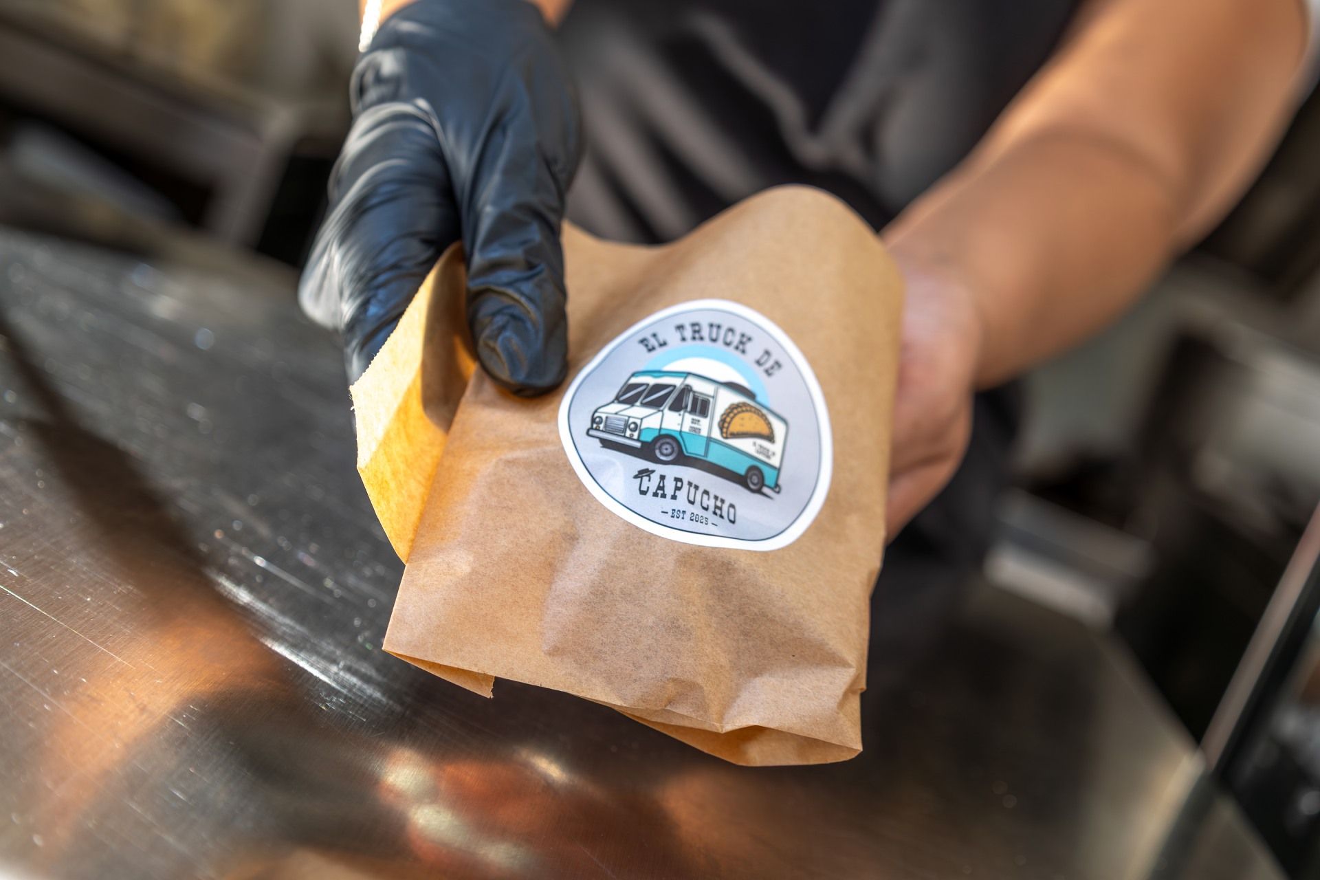 Eine Hand in einem schwarzen Handschuh hält eine braune Papiertüte mit einem Aufkleber mit dem Logo eines Imbisswagens.