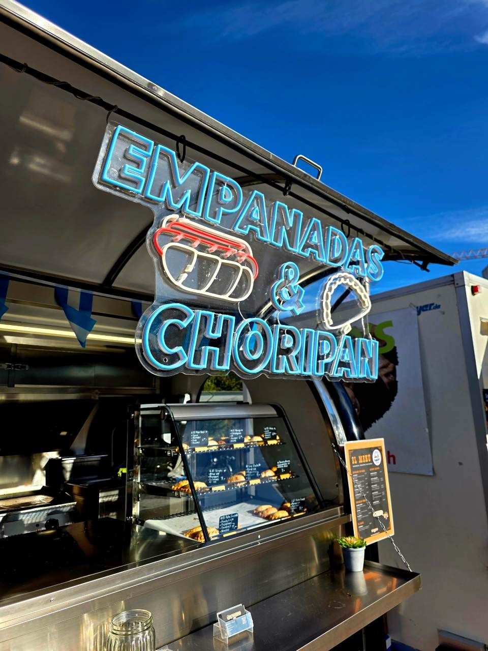 Ein Imbisswagen serviert Empanadas und Choripan; über einer Vitrine mit Speisen unter blauem Himmel prangt ein helles Neonschild.