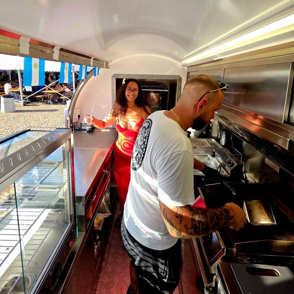 Ein tätowierter Koch arbeitet an einem Grill in einem mobilen Imbisswagen, während eine lächelnde Frau in einem roten Kleid daneben steht.