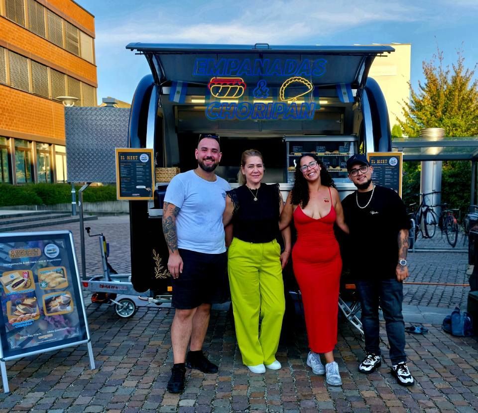 Vier Personen stehen lächelnd vor einem Imbisswagen und posieren für ein Foto auf einem gepflasterten Platz im Freien.