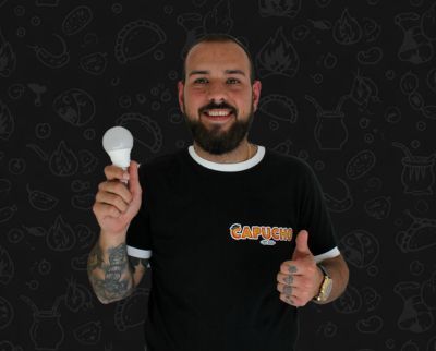 Eine Person mit Bart, die ein schwarzes Hemd trägt, hält eine Glühbirne und zeigt vor einem dunklen, gemusterten Hintergrund den Daumen nach oben.