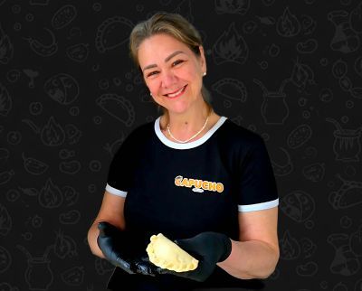 Eine lächelnde Person in schwarzem T-Shirt und Handschuhen hält eine frisch zubereitete Empanada vor einem gemusterten schwarzen Hintergrund.