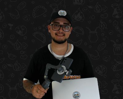 Eine Person mit Kappe, Brille und Kette hält ein Handy-Gimbal und einen Laptop vor einem gemusterten schwarzen Hintergrund.