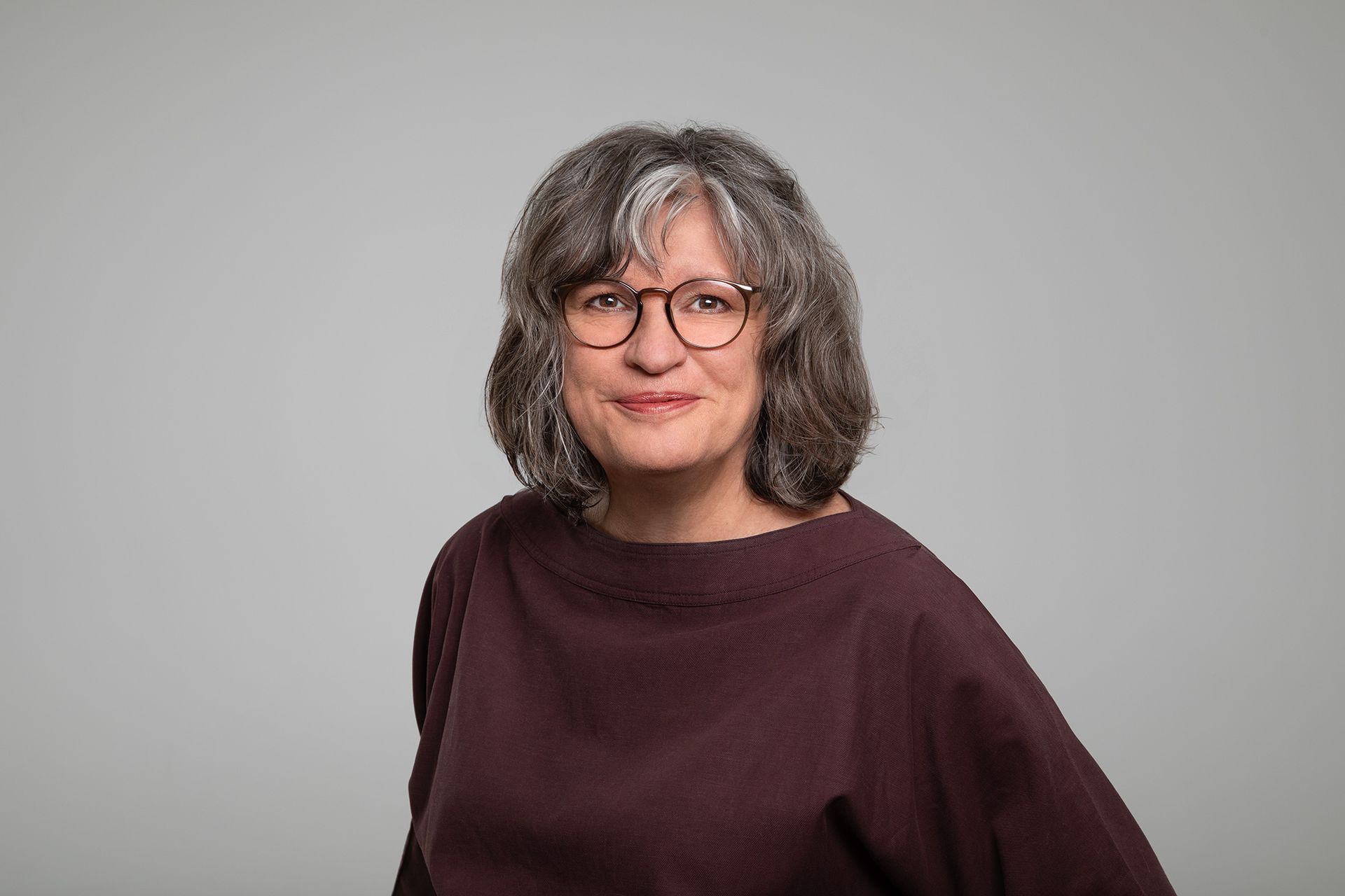Porträt einer Frau mit grauem, lockigem Haar und Brille vor einem neutralen grauen Hintergrund.