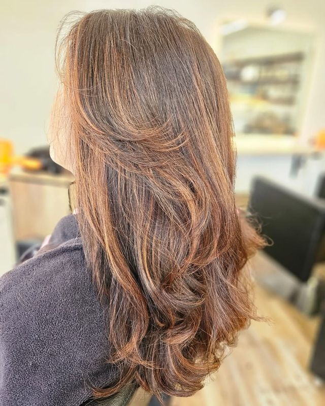 Cabello largo, ondulado, castaño oscuro con reflejos más claros y puntas capeadas, visto desde atrás en un salón de belleza.