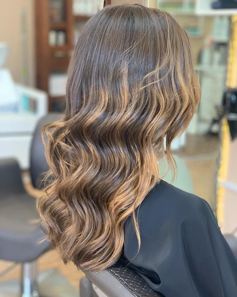 Cabello largo, ondulado y castaño oscuro con mechas balayage rubio miel cálido, visto desde atrás en un salón de belleza.