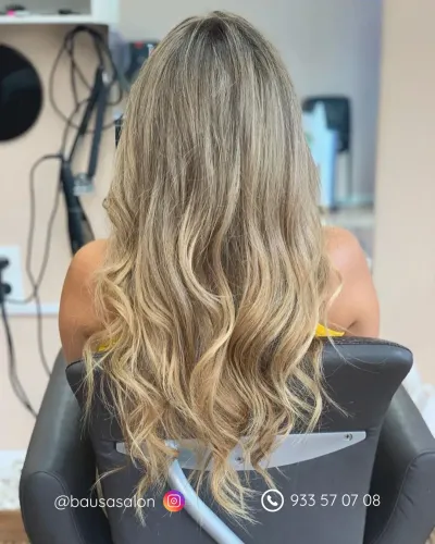 Una persona con cabello rubio largo, ondulado y con capas, vista de espaldas, sentada en una silla.