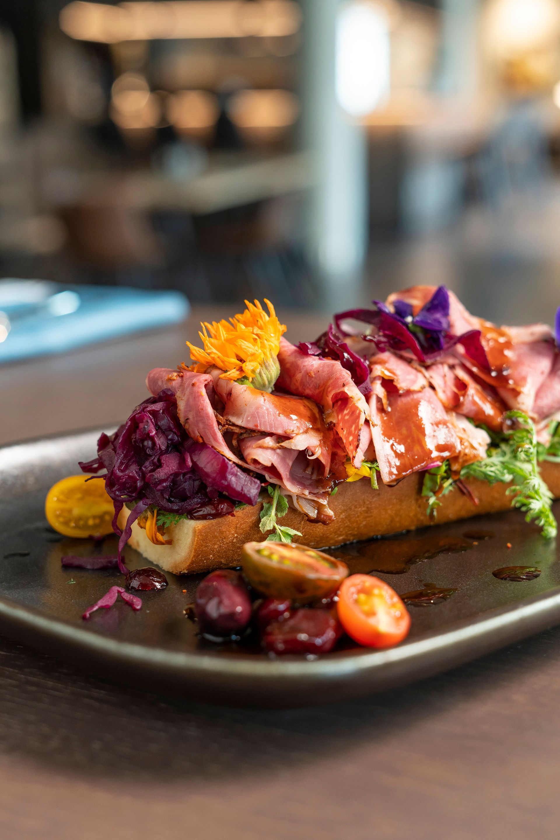 Ein Gourmet-Sandwich mit Roastbeef, Rotkohl, Kirschtomaten und gelben Blüten auf einem dunklen, rechteckigen Teller.