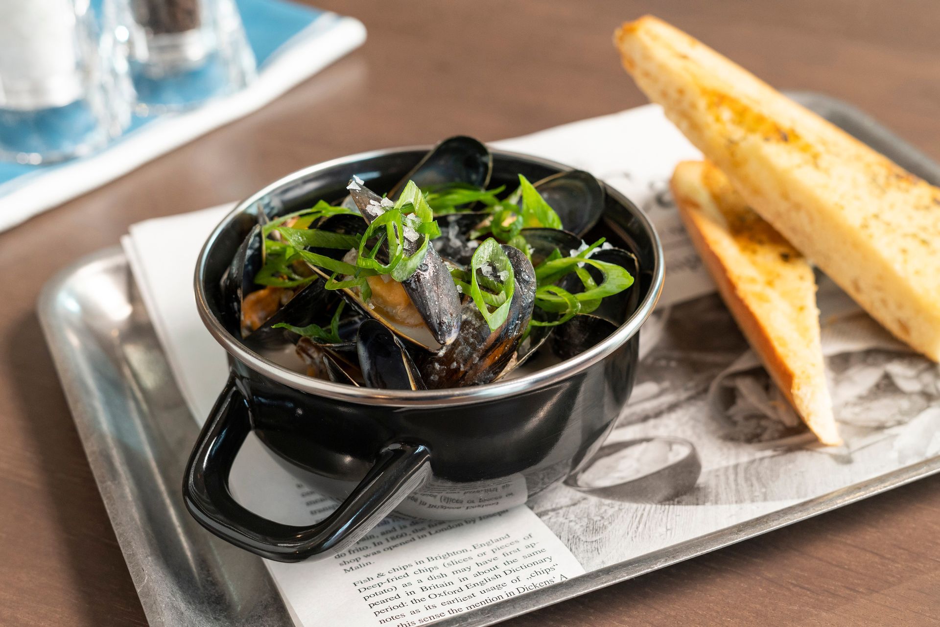 Ein Metalltopf gefüllt mit Muscheln, garniert mit grünen Kräutern, serviert mit zwei Scheiben geröstetem Brot auf einem Tablett mit Zeitungspapier.